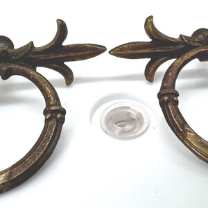 Könnte beinhalten: Zwei Schubladengriffe aus Messing im antiken Stil mit einem kreisförmigen Ringgriff, der an einer dekorativen horizontalen Halterung mit blattartigen Akzenten hängt. Das Metall hat ein verwittertes, dunkelgoldenes Finish mit Patina.
