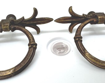 May include: Two antique-style brass drawer pulls featuring a circular ring handle suspended from a decorative horizontal mount with leaf-like accents. The metal has a weathered, dark gold finish with visible patina and textured detailing on the rings.
