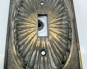 May include: A rectangular metal light switch plate featuring a central oval medallion with a fluted, sunburst-like design. The plate has a dark, aged bronze finish with highlights of gold. It includes two screw holes and a central rectangular cutout.