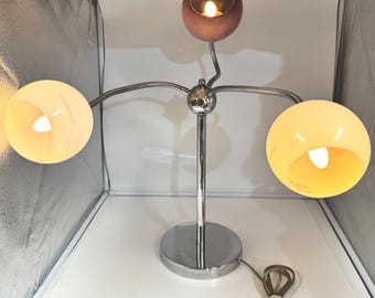 May include: A three-light table lamp featuring a central chrome pole and base with three adjustable, flexible chrome arms. Each arm holds a spherical glass shade in distinct colors: one pale pink, one soft yellow, and one light cream, each with a bulb.
