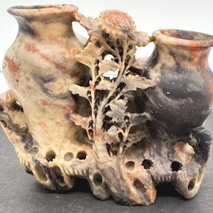 May include: A carved stone vase with two openings, connected by a floral and leaf design. The vase has a natural colour palette of beige, brown, and black. The intricate carving adds detail to the decorative piece.