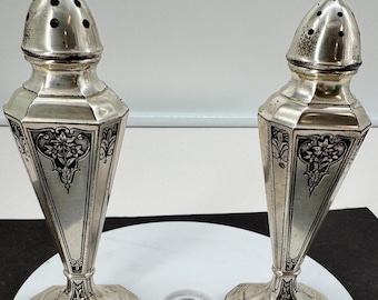 May include: A pair of silver-toned metal salt and pepper shakers with a tall, tapered hexagonal body. Each shaker features a removable domed cap with small circular holes and intricate black floral engravings on the side panels and base.