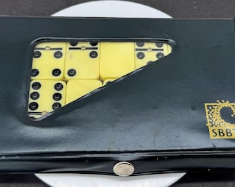 May include: A black rectangular case with a triangular cutout window revealing yellow dominoes with black pips inside. The case features a gold logo with the text SBBT and a single silver-colored snap button closure at the bottom edge.