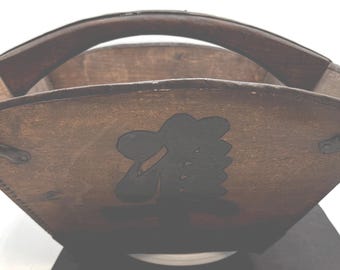 May include: A wooden basket with a curved handle. The basket is brown with a dark emblem on the front. The handle is also brown and appears to be made of wood. The basket has a square shape.