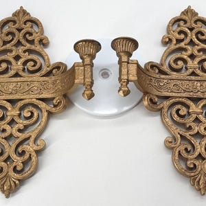 Puede incluir: Par de apliques de pared dorados y ornamentados. Cada aplique presenta un diseño decorativo con volutas y dos candelabros. Los apliques tienen una barra horizontal curvada con tallas detalladas. El estilo general es vintage y elegante.