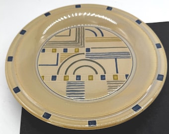 May include: A beige ceramic plate with a geometric design. The plate has a wide rim with dark blue square accents. The center features a pattern of blue, yellow, and gray lines and arcs on a cream background. A decorative piece for the kitchen.