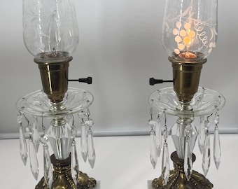 May include: Two antique table lamps with clear glass shades etched with floral designs. The lamps have brass fixtures, crystal accents, and sit on marble bases. The lamps are lit, casting a warm glow.