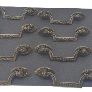 Puede incluir: Un juego de ocho tiradores de cajón de latón de estilo antiguo con un diseño de asa curva y patrones florales en relieve en el mango y las placas de montaje, acompañado de un pequeño montón de tornillos de montaje de metal plateado.