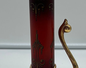 May include: A gold-toned candle holder with a red glass center. The holder has a decorative design with gold accents and a curved handle.