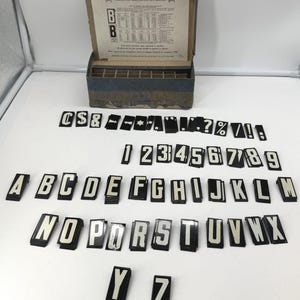 May include: A vintage typesetting kit with a wooden box containing individual letterpress letters and numbers. The letters are black with white lettering. The box has a price list for additional letters.