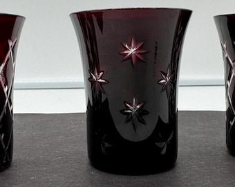 May include: Three dark red glass tumblers with cut glass designs. The tumblers have different patterns, one with a diamond pattern, one with a star pattern, and one with a combination of both.