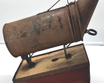 May include: A vintage metal bee smoker featuring a cylindrical, rusted brown canister with a tapered nozzle and a metal handle. The canister is mounted onto a wooden and red bellows base, which includes a small circular hole in the wood surface.