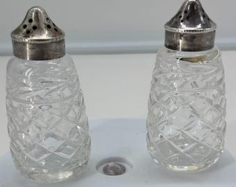 May include: A pair of clear glass salt and pepper shakers featuring a diamond-cut pattern on the body and rounded silver-toned metal caps with small perforated holes at the top for dispensing contents. The shakers have a tapered cylindrical shape.