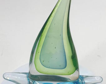 May include: A decorative glass sculpture in the shape of a sailboat. The sail is a layered design of green and blue hues, while the base features a swirling blue and white pattern. The sculpture is transparent, allowing light to pass through.