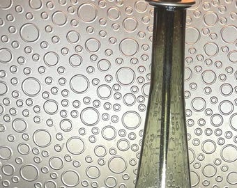 May include: A vintage table lamp with a tall, olive green glass base and a brass-colored metal base and accents. The lamp has a clear lightbulb and a brass-colored metal shade holder. The background is a textured silver wall.