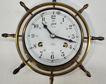 May include: A brass ship's wheel clock with a white face, black hands, and Arabic numerals. The dial features the text Schatz, Ships Bell, 8 Day, 7 Jewels, and Made in Germany. It includes an on-off switch at the top and two winding holes.