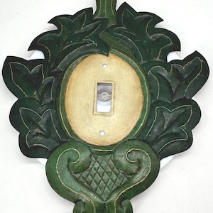May include: A decorative light switch plate featuring a central oval cream-colored panel surrounded by dark green carved leaf motifs and a bottom section with a diamond-patterned texture. The plate has a rectangular cutout for a toggle switch.