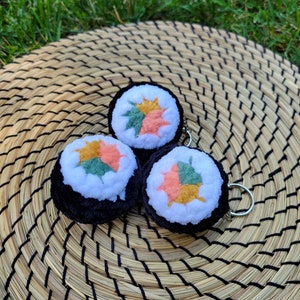 May include: Three plush sushi roll keychains with black seaweed, white rice, and colorful leaf fillings. The keychains are arranged on a woven mat, with green grass in the background. Each sushi roll is approximately 2 inches in diameter.