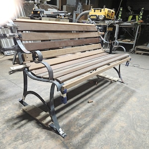 May include: A wooden bench with a dark metal frame. The bench has multiple wooden slats for the seat and backrest. The metal frame has decorative curved armrests. The bench is in a workshop setting.