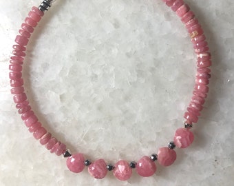 May include: A pink gemstone bracelet with a silver clasp. The bracelet features small, round pink beads and larger, faceted pink beads. The clasp is a toggle style. The bracelet is displayed on a white surface.