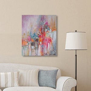 May include: Abstract painting with pink, orange, and blue colors on a white canvas. The painting is hanging on a wall in a living room setting with a couch and a floor lamp.