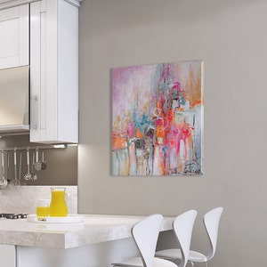 Op de afbeelding: Abstract schilderij met roze, oranje, blauwe en witte kleuren. Het schilderij hangt aan een muur in een keuken met witte kasten en een wit aanrecht.