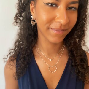 Puede incluir: Una mujer lleva un collar de plata con un colgante de coraz&oacute;n y un peque&ntilde;o colgante de perla. Tambi&eacute;n lleva pendientes dorados. La mujer lleva una blusa azul marino.