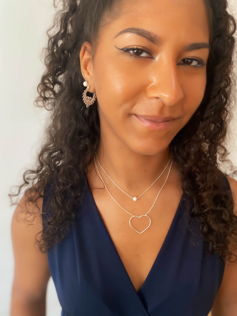 Puede incluir: Una mujer lleva un collar de plata con un colgante de coraz&oacute;n y un peque&ntilde;o colgante de perla. Tambi&eacute;n lleva pendientes dorados. La mujer lleva una blusa azul marino.