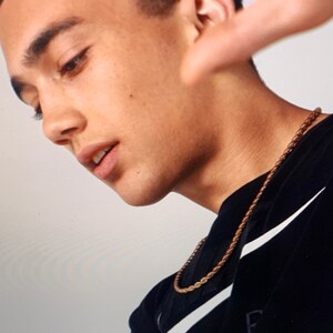 May include: A young person wearing a black shirt with a white Nike swoosh and a gold chain necklace. The person is looking down and has their hand raised to their face.