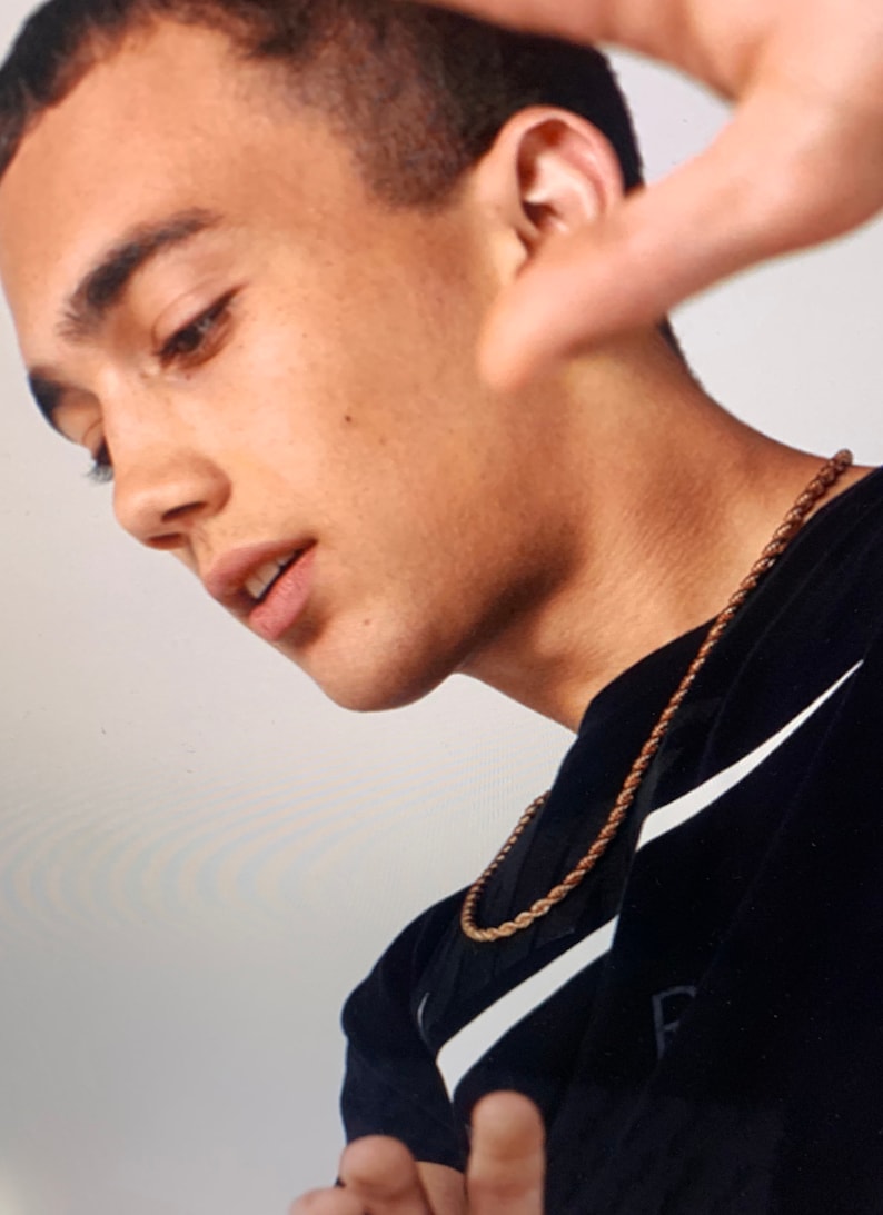 May include: A young person wearing a black shirt with a white Nike swoosh and a gold chain necklace. The person is looking down and has their hand raised to their face.