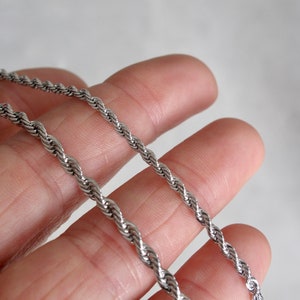 May include: Two silver rope chains of different thicknesses, laid out on a hand.
