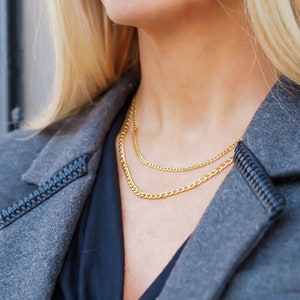May include: A woman wearing a gray jacket with a black shirt underneath. She is wearing two gold chain necklaces.