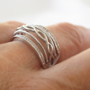 Puede incluir: Un anillo plateado con un diseño intrincado. El anillo presenta múltiples bandas, algunas adornadas con pequeñas piedras transparentes, y otras con un patrón tejido. El anillo se muestra en el dedo de una persona, destacando sus detalles.
