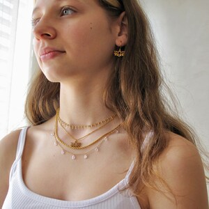 May include: A woman wearing a white vest top with a layered gold necklace featuring a small pendant. The necklace has a delicate chain with small, clear beads.