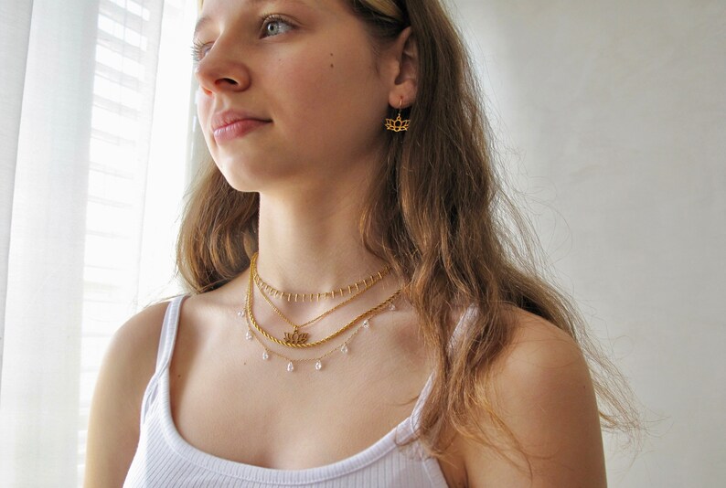 May include: A woman wearing a white vest top with a layered gold necklace featuring a small pendant. The necklace has a delicate chain with small, clear beads.