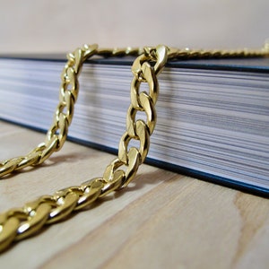 May include: A gold chain necklace on a black book. The chain is thick and has a curb link design.