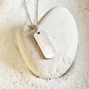 Peut inclure: Collier en argent avec un pendentif rectangulaire. Le pendentif porte le nom "Alara" gravé et un motif de main de Hamsa. Le collier est sur une pierre blanche en forme de cœur.