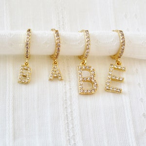 Peut inclure: Quatre boucles d'oreilles créoles dorées avec des breloques en forme de lettres pendantes. Les breloques sont en or et ornées de strass transparents. Les lettres sont B, A, B et E.