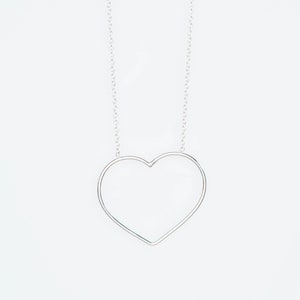 Puede incluir: Un collar plateado con un colgante en forma de coraz&oacute;n. El colgante es un simple contorno de coraz&oacute;n, suspendido de una fina cadena. El collar se muestra sobre un fondo blanco liso.