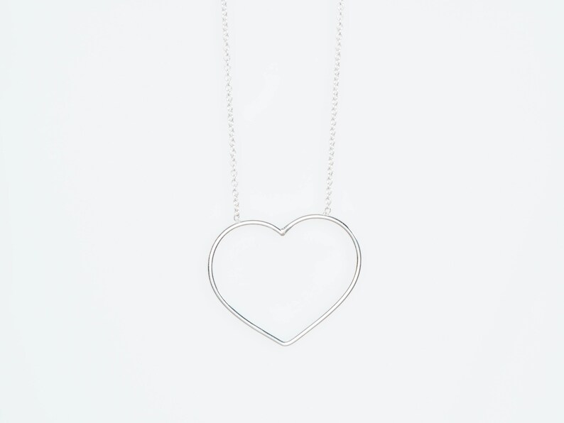 Puede incluir: Un collar plateado con un colgante en forma de coraz&oacute;n. El colgante es un simple contorno de coraz&oacute;n, suspendido de una fina cadena. El collar se muestra sobre un fondo blanco liso.