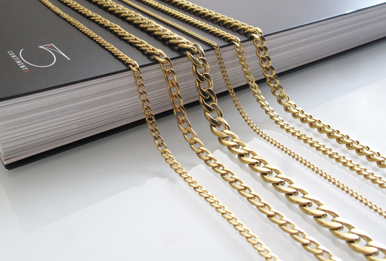 May include: A set of seven gold chains of varying widths and lengths. The chains are arranged in a fan shape on a black book with the word "CONTINENT" printed on the cover.