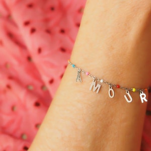 Peut inclure: Un bracelet à breloques argenté avec le mot "AMOUR" écrit en breloques argentées. Le bracelet est orné de perles colorées.