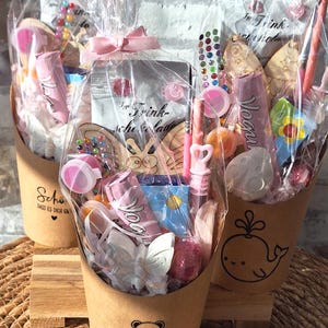 Könnte beinhalten: Drei Geschenk-Körbe gefüllt mit verschiedenen Artikeln, verpackt in durchsichtigem Zellophan und mit rosa Bändern gebunden. Die Körbe sind aus braunem Papier und mit einem Bären, einem Wal und einem Schmetterling verziert. Sie enthalten Süßigkeiten, Lipgloss und Holzzuschnitte.