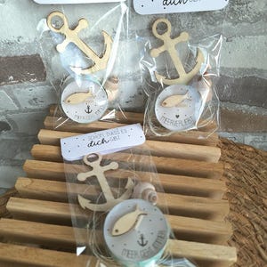 Könnte beinhalten: Drei kleine Geschenkbeutel mit maritimen Dekorationen. Jeder Beutel enthält einen Holzanker, eine runde Scheibe mit einer Fischillustration und eine Muschel. Der Text "SCHON, DASS ES dich GIBT" ist oben auf jedem Beutel aufgedruckt.