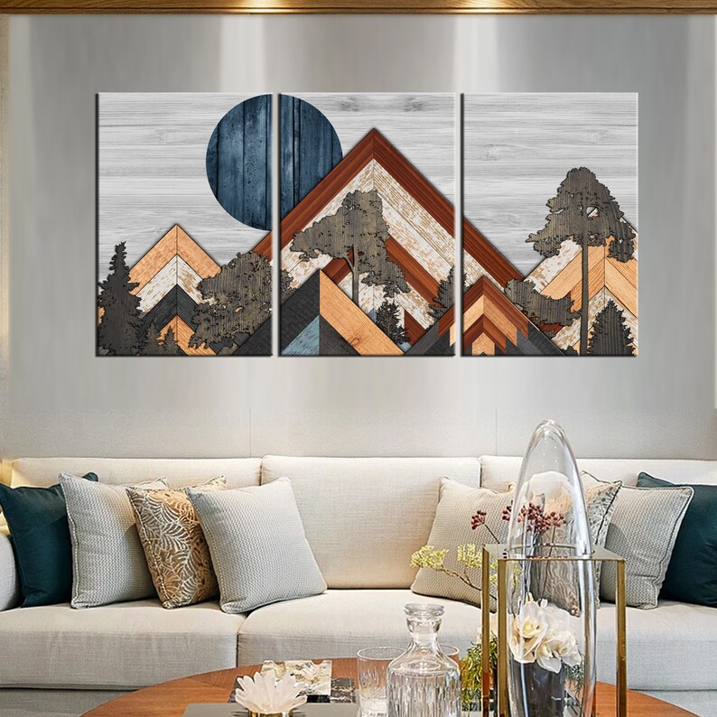 May include: A three-panel wall art piece featuring a mountain landscape design. The artwork uses geometric shapes and wood textures in shades of brown, orange, blue, and gray. A large blue circle represents the sun or moon. Trees are also included in the design.