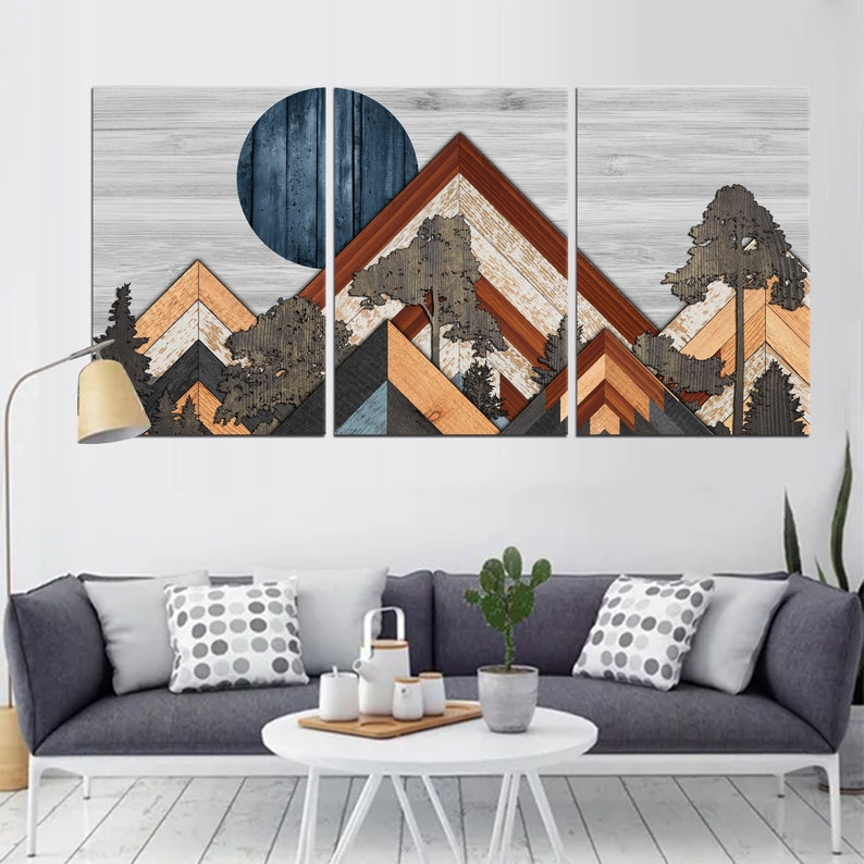 May include: A three-panel wooden wall art piece depicting a mountain landscape. The artwork features geometric mountain shapes in shades of brown, orange, gray, and blue, with a dark blue circle representing the sun or moon. Trees are also incorporated into the design.