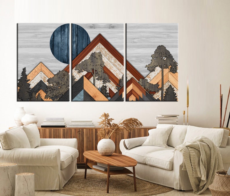 May include: A three-panel wooden wall art piece featuring a mountain landscape design. The artwork incorporates geometric shapes and natural wood tones, with a blue circle representing the sun or moon. Trees are also included in the design.