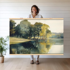 Puede incluir: Una pintura de acuarela de una escena de lago con árboles reflejados en el agua. Los árboles son una mezcla de verde y amarillo, y el agua es de un azul claro. La pintura está enmarcada en un marco de madera.