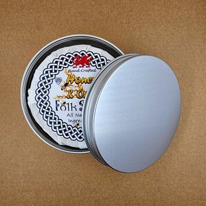 May include: A silver tin with a white label that reads "Hand Crafted Honey & Oat Folk Soap All Natural Ingredients". The tin is open and the lid is sitting next to it.
