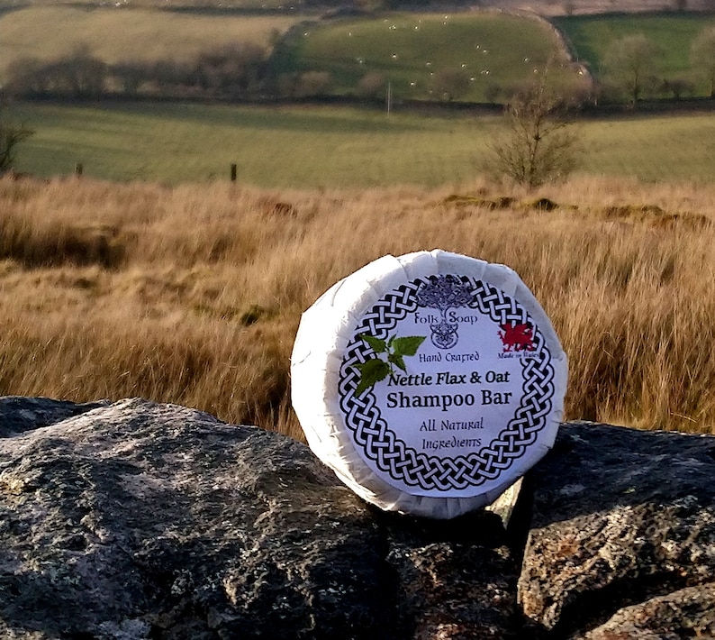 May include: A white round bar of soap with a black and white Celtic knot design. The label reads "Folk Soap Hand Crafted Nettle Flax & Oat Shampoo Bar All Natural Ingredients".